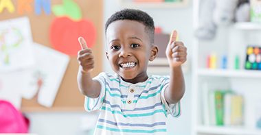 An adorable preschool age little boy smiles for the camera as he stands in his preschool classroom and gives a thumbs up.  He loves school!